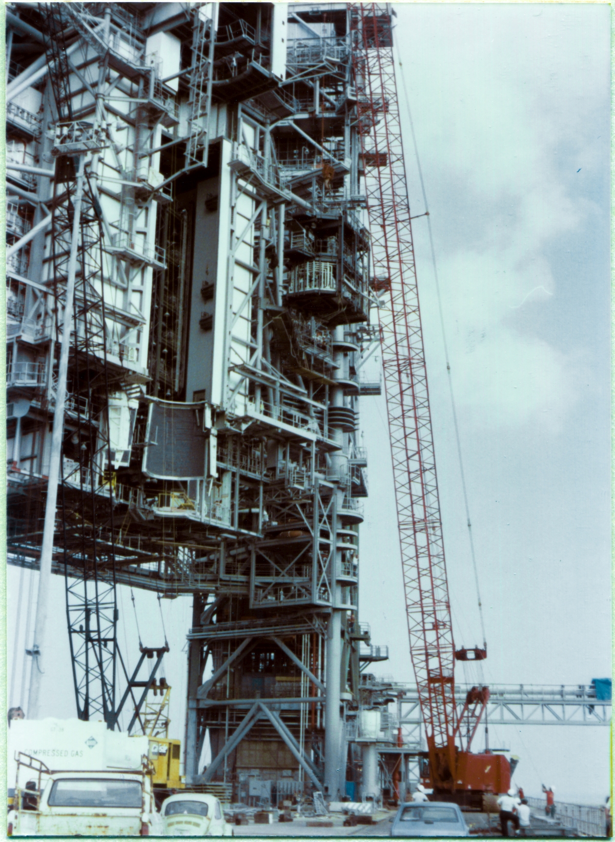 Image 093. At Space Shuttle Launch Complex 39-B, Kennedy Space Center, Florida, the Orbiter Mid-Body Umbilical Unit Lift has proceeded to the point where the OMBUU has reached its final elevation, and now the crane operator is booming right, ever so slowly, ever so carefully, bringing the OMBUU closer and closer to where it will rest against the face of the Rotating Service Structure and be bolted permanently into place. The criticality of the work the two Union Ironworkers working for Ivey Steel, down on the ground, work-gloved hands gripping the tag lines, fine-positioning the OMBUU, has stepped up a couple of notches, and you can see them both, over by the Flame Trench, putting their backs into it, with the tag lines clearly in significant tension, keeping the OMBUU from bashing into anything up on the RSS as it slowly, creepingly, closes in to a steel-on-steel position. Additional personnel, above and beyond the gang of ironworkers who will be doing the actual connecting, can be seen at the end of the OMBUU Access Catwalk at Elevation 163'-9”, watching things closely. Quality, Safety, Craft Labor, Management, Engineering, Contractor, Structural, Mechanical, Electrical, NASA-direct, and more, all had very good and sufficient reason to be eyes-on as close as possible, at this stage of the Lift. Photo by James MacLaren.